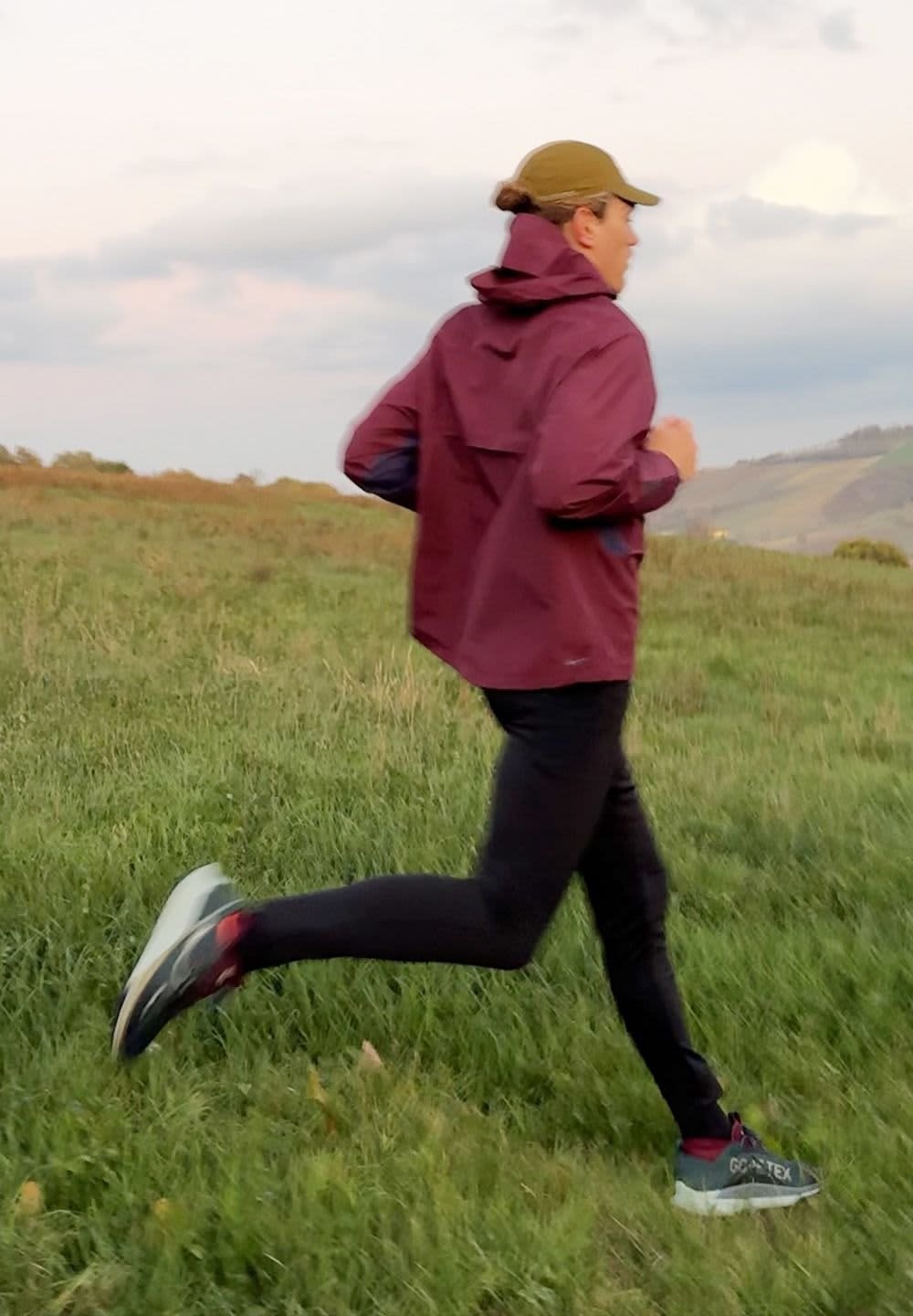 Maroon waterproof jacket with a hood, black pants, and grey running shoes with red accents. Runner in a grassy field during twilight.
