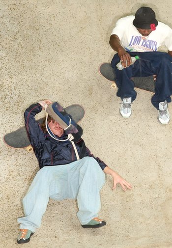 Twee skateboarders in een skatepark. De een ligt op de grond en draagt een blauwe jas en lichtblauwe broek. De ander zit op een skateboard en houdt een fles vast.