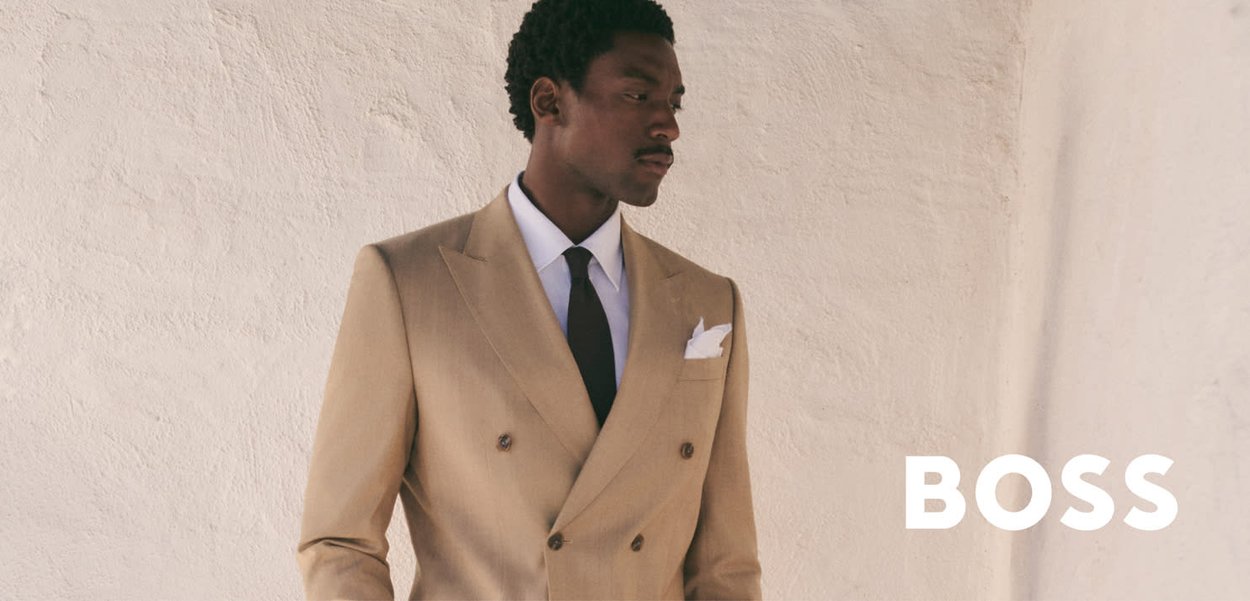 Hombre con traje cruzado color tostado, camisa blanca y corbata negra, de pie contra una pared texturizada beige con la palabra BOSS.
