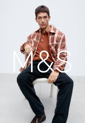 Rote und weiß karierte Jacke über einem rostfarbenen Pullover, kombiniert mit dunklen Hosen und Lederschuhen, sitzend auf einem Stuhl vor einfachem Hintergrund.