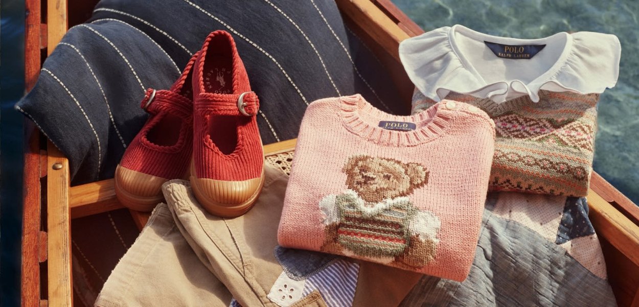 Gefaltete Kinderkleidung, darunter ein rosa Pullover mit Teddybär, ein gemusterter Pullover mit weißem Kragen, beige Hosen, rote Schuhe und Kissen in einem Holzboot.