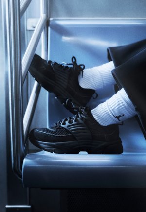 Jambes en pantalon noir et chaussettes blanches portant des baskets noires reposant sur un siège bleu clair de bus ou de train avec des rampes métalliques.