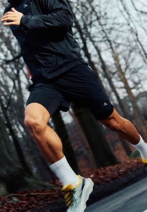 Läufer trägt schwarze Shorts, schwarze Jacke, weiße Socken und graue Turnschuhe und joggt auf einem Waldweg mit kahlen Bäumen im Hintergrund.