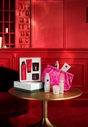 Set di prodotti di bellezza in una scatola rossa, che include una pochette rosa trapuntata, due candele e un diffusore, esposti su un tavolo rotondo dorato.
