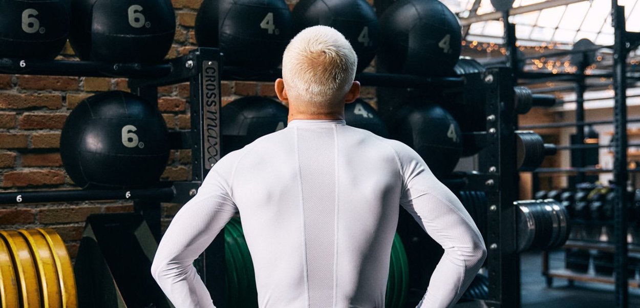 Osoba o krótkich blond włosach ubrana w biały strój sportowy stoi twarzą do stojaków z piłkami lekarskimi i talerzami obciążeniowymi na siłowni.