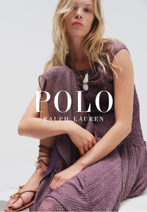 Mujer rubia con vestido sin mangas de patrón morado, sandalias marrones y un collar blanco con colgante, sentada con los brazos cruzados sobre las rodillas.