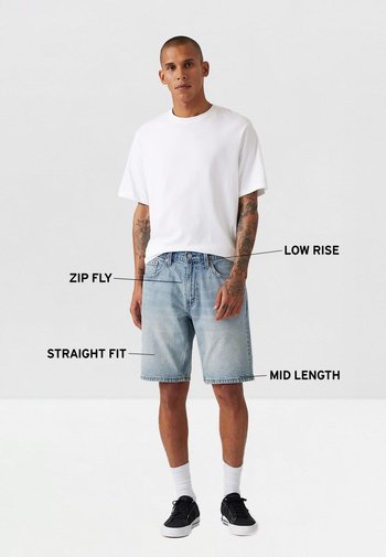 Camiseta blanca de manga corta, pantalones cortos de mezclilla azul claro con cremallera, de tiro bajo, corte recto, de longitud media, combinados con zapatillas negras y calcetines blancos.