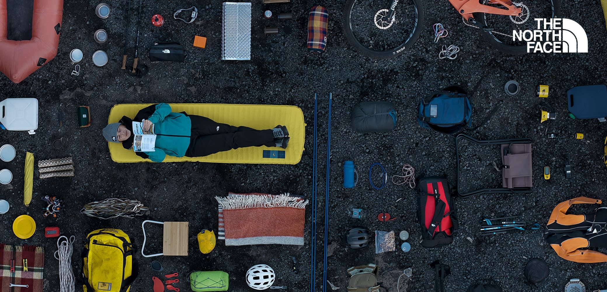 Materassino giallo con persona che legge, circondata da attrezzature da campeggio: tende, zaini, pentole e attrezzi su sfondo di ghiaia scura.