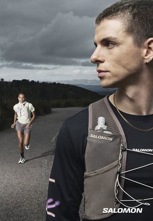 Uomo che indossa un gilet da corsa Salomon che guarda di lato, donna che corre su una strada sullo sfondo sotto un cielo nuvoloso.