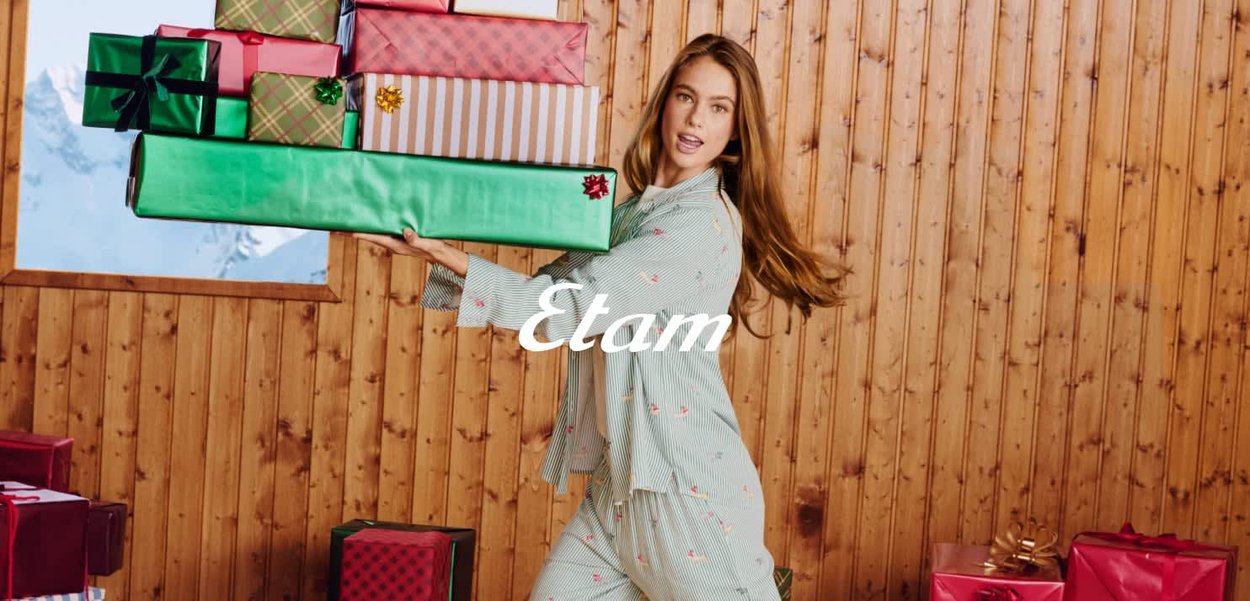 Modèle en pyjama rayé tenant une grande boîte cadeau verte avec un ruban, entouré de cadeaux colorés emballés, fond en bois.