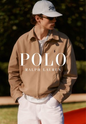 Joven con chaqueta marrón claro, pantalones blancos, gorra blanca de béisbol y gafas de sol, de pie al aire libre con las manos en los bolsillos.