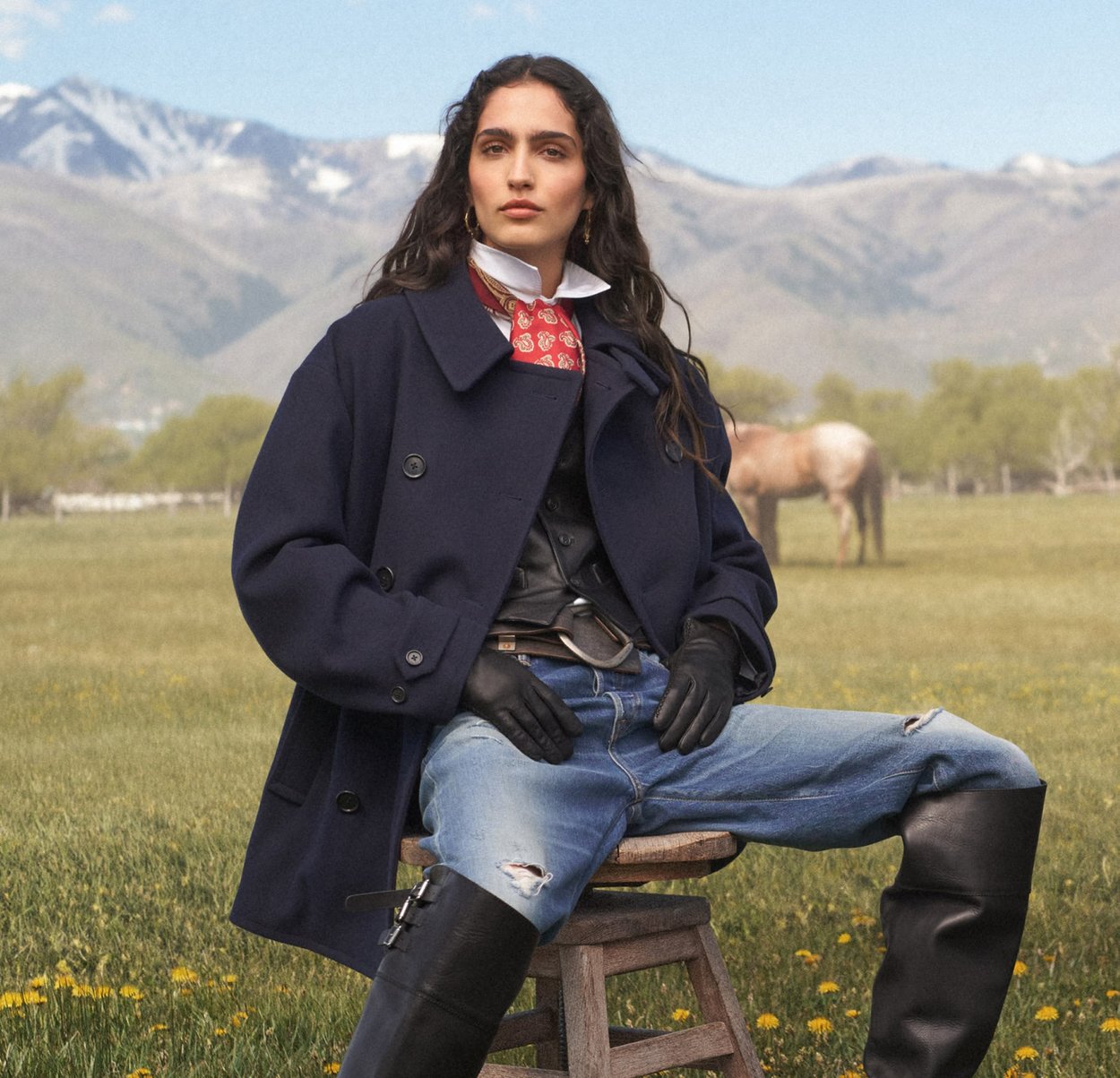 Cappotto di lana blu navy con colletto ampio, indossato sopra un gilet nero, abbinato a jeans blu strappati e stivali neri di pelle alti. Accento con sciarpa rossa.