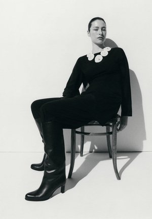 Woman wearing black outfit with large white floral necklace and black knee-high boots, sitting on wooden chair against white background.