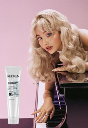 Redken Acidic Bonding Concentrate leave-in behandling i en hvid tube med blå tekst. Model med langt, bølget blondt hår nær et klaver.