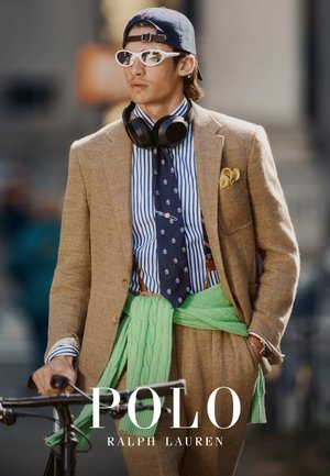 Jonge man die een beige pak draagt, een gestreept overhemd, een stropdas met patroon, witte zonnebril, een pet achterstevoren, koptelefoon en een groene trui om zijn middel, terwijl hij een fiets vasthoudt.