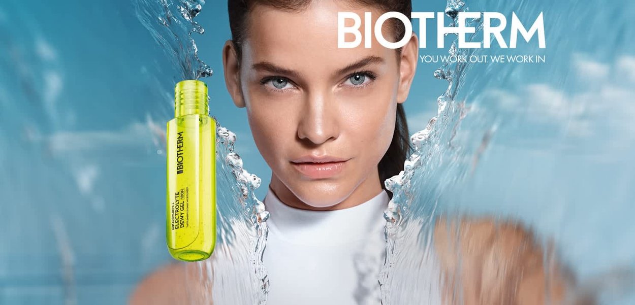 Frau mit nassen Haaren blickt direkt nach vorne, während Wasser spritzt und ein Biotherm-Hautpflegeprodukt auf blauem Hintergrund in der Nähe schwebt.