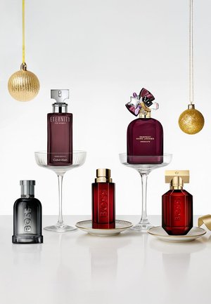 Five perfume bottles with varying shapes and colors: dark purple, red, and black glass. Gold accents and holiday ornaments in the background.