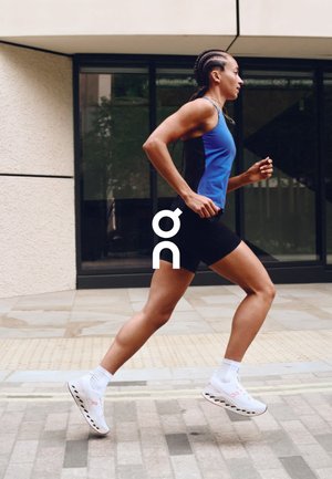 Running shoes with a white and black sole, mesh upper in blue and black. Model in activewear jogging on a textured outdoor path.
