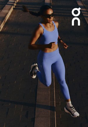 Mujer corriendo al aire libre por un camino pavimentado con ropa deportiva azul, zapatillas blancas para correr y gafas de sol oscuras bajo la luz del sol.