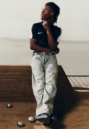 Maglietta Nike in bianco e nero, jeans baggy azzurri, cintura con borchie, in piedi su terreno sabbioso, con palline metalliche visibili nel primo piano.