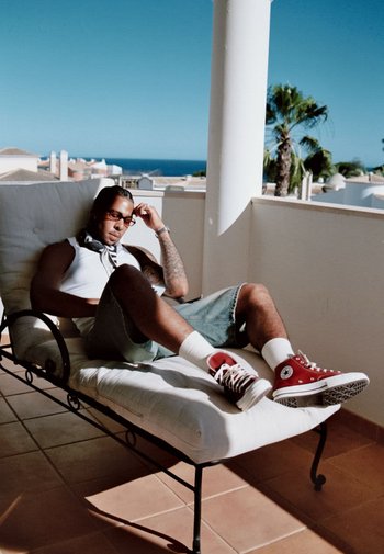Red canvas high-top sneakers, white ankle socks, light blue denim shorts, and a white tank top; seated on a white cushioned lounge chair.