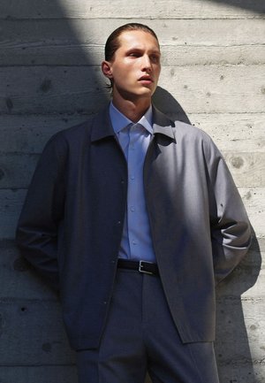 Giacca leggera grigia sopra una camicia azzurro chiaro con bottoni, abbinata a pantaloni grigi. Tessuti morbidi, design minimalista, dettagli dei bottoni visibili.