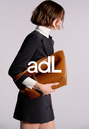 Bruine leren handtas met een gestructureerd ontwerp, met een contrasterende lichtbruine band. Model draagt een zwarte outfit met witte accenten en houdt de tas vast.