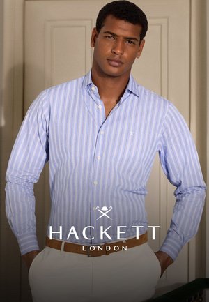 Man in een blauw-wit gestreepte overhemd, ingestopt in witte broek met een beige riem, staande binnen tegen een wand met panelen.