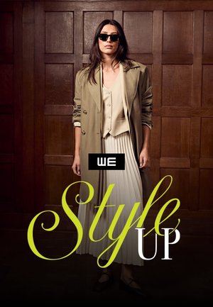 Vrouw met zonnebril, beige trenchcoat, dichtgeknoopte vest en geplooide rok, staand tegen een houten wand met panelen.