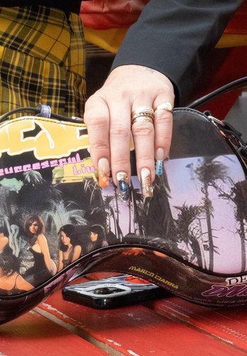 Zwarte handtas met kleurrijke afbeeldingen van vrouwen en palmbomen. Heeft een gebogen vorm en een glanzende afwerking. Een hand met nagelkunst leunt erop.