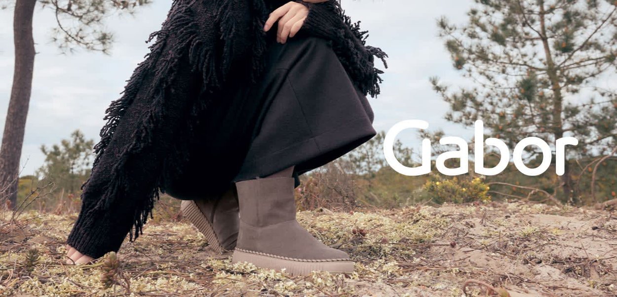 Bottines en daim gris à bout rond, semelle contrastée et coutures décoratives ; portées avec un long vêtement noir dans un cadre naturel.