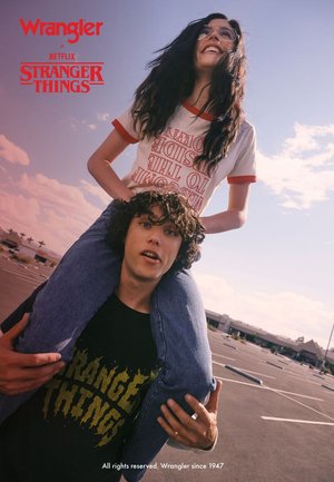 Zwei junge Erwachsene posieren im Freien: Einer trägt ein weißes T-Shirt mit roten Akzenten, der andere ein schwarzes "Stranger Things"-Shirt. Helles Sonnenlicht.