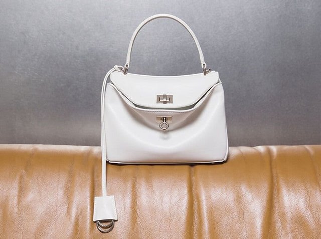 Sac à main en cuir blanc avec une poignée supérieure et une fermeture à rabat, posé sur un canapé en cuir marron contre un mur gris.