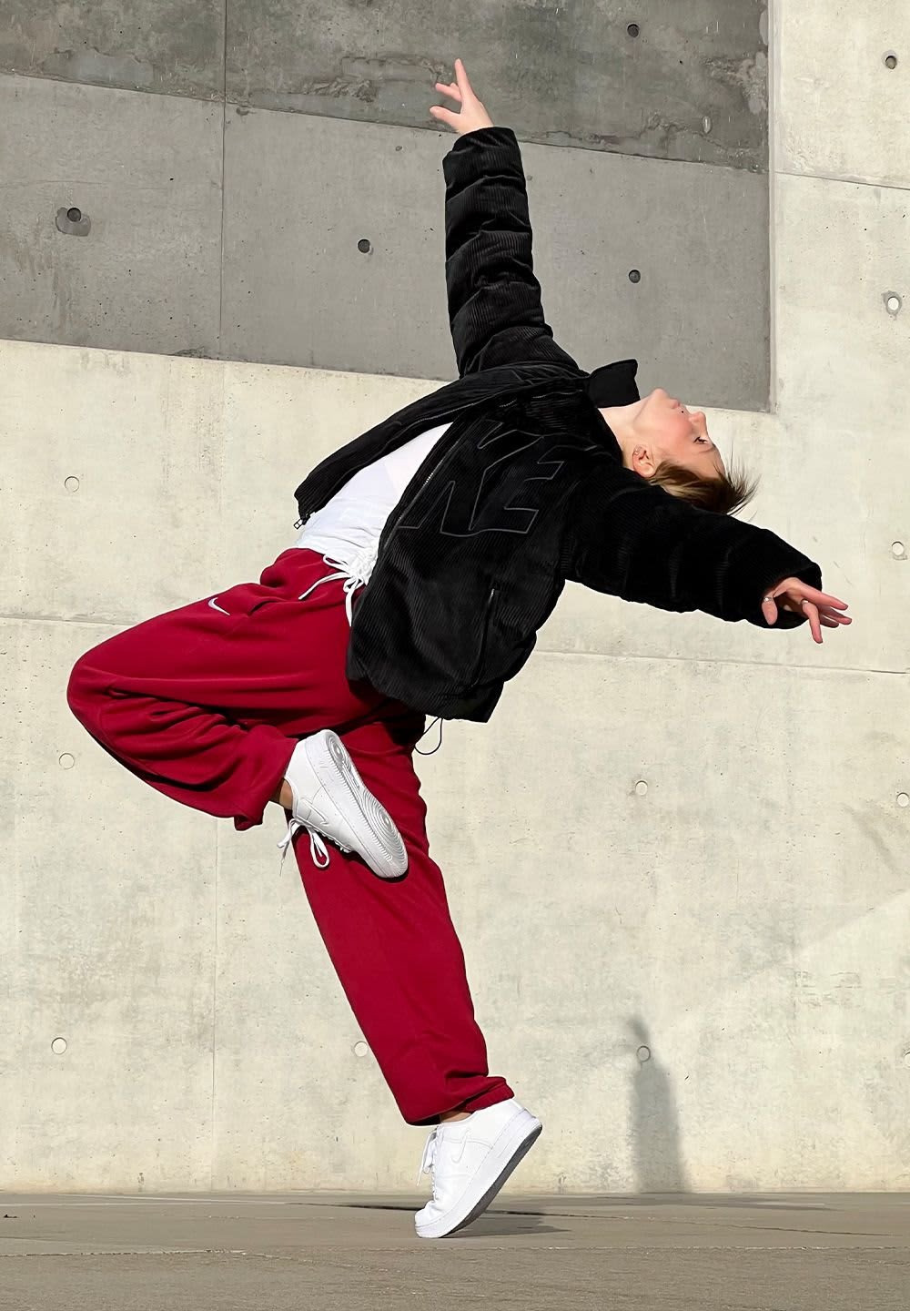 Black corduroy jacket with a large logo, white t-shirt, red sweatpants, and white sneakers, posed dynamically against a concrete backdrop.