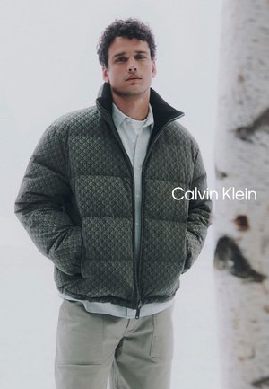 Padded jacket in olive green with a geometric pattern, high collar, and zip closure, worn over a white shirt, set against a light background.