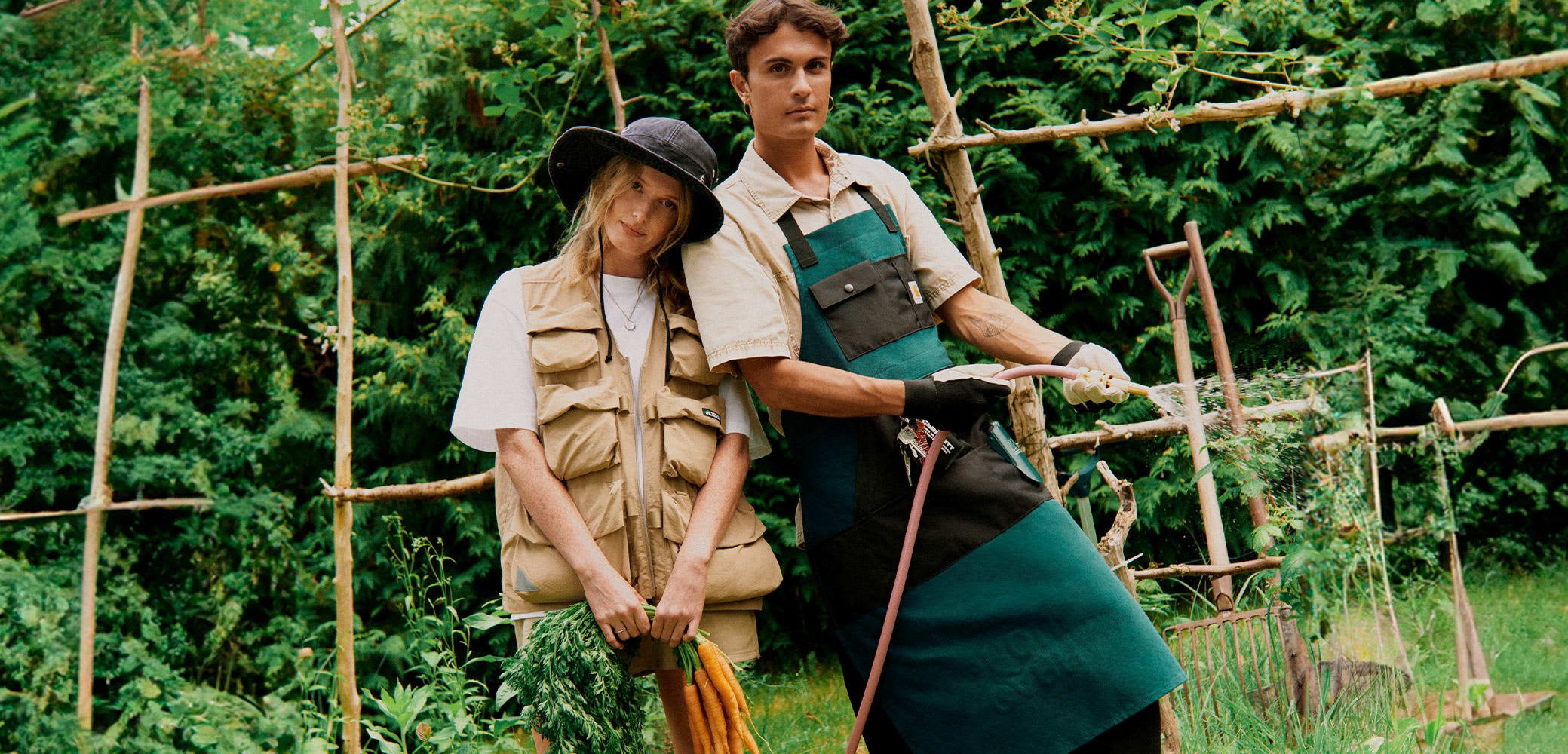 Two individuals stand together in a garden. One wears a beige utility vest and holds green foliage. The other wears a teal apron and uses a hose.