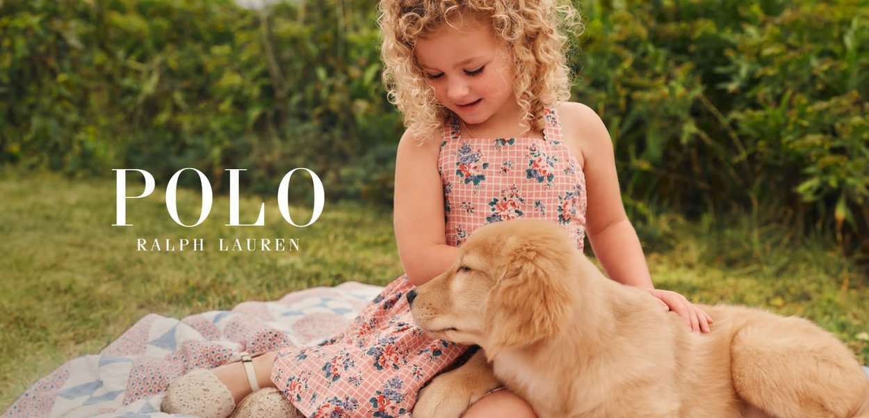 Niña joven con vestido de flores sentada en una manta al aire libre, acariciando suavemente a un golden retriever que yace a su lado.