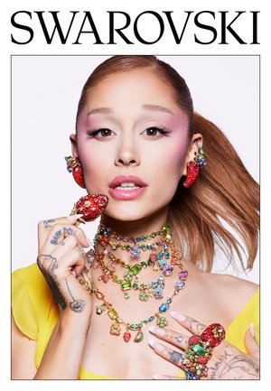 Femme aux cheveux roux portant des colliers en pierres précieuses colorées, des boucles d'oreilles et une bague en forme de fraise, ainsi qu'un haut jaune, tenant un accessoire en forme de fraise.