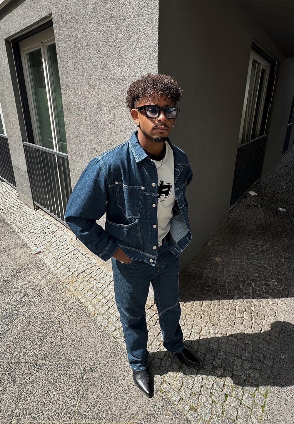 Mann mit dunkler Sonnenbrille, Jeansjacke und Jeans, weißem Grafik-T-Shirt, der auf einem Kopfsteinpflaster in der Nähe eines Gebäudes im Sonnenlicht steht.