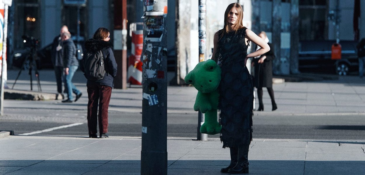 Woman in a sleeveless black dress with circular textures holds a large green teddy bear, standing on a city street with pedestrians in the background.