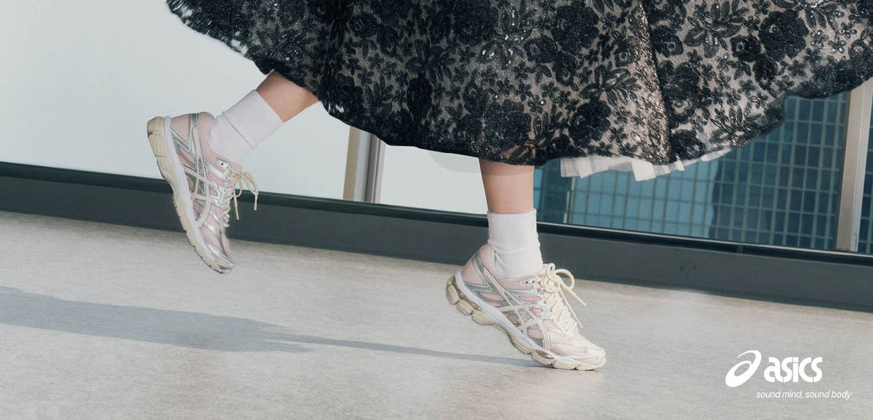 Osoba nosząca białe skarpetki i srebrne buty sportowe Asics w połowie kroku, pod czarną koronkową spódnicą, w pomieszczeniu blisko okna z widokiem na miejskie budynki.