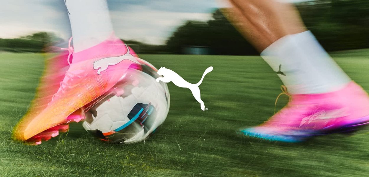 Persona con botas Puma rosas regateando un balón de fútbol blanco sobre hierba verde con efecto de movimiento.