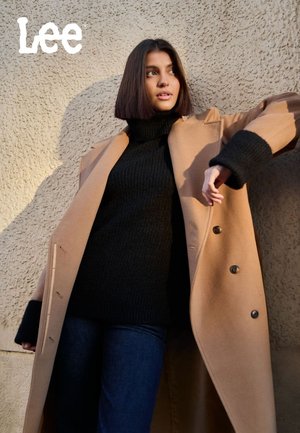 Kamel overcoat med svart ribbestrikket turtleneck, mørkeblå jeans. Har store knapper og et strukturert design. Teksturert veggbakgrunn.