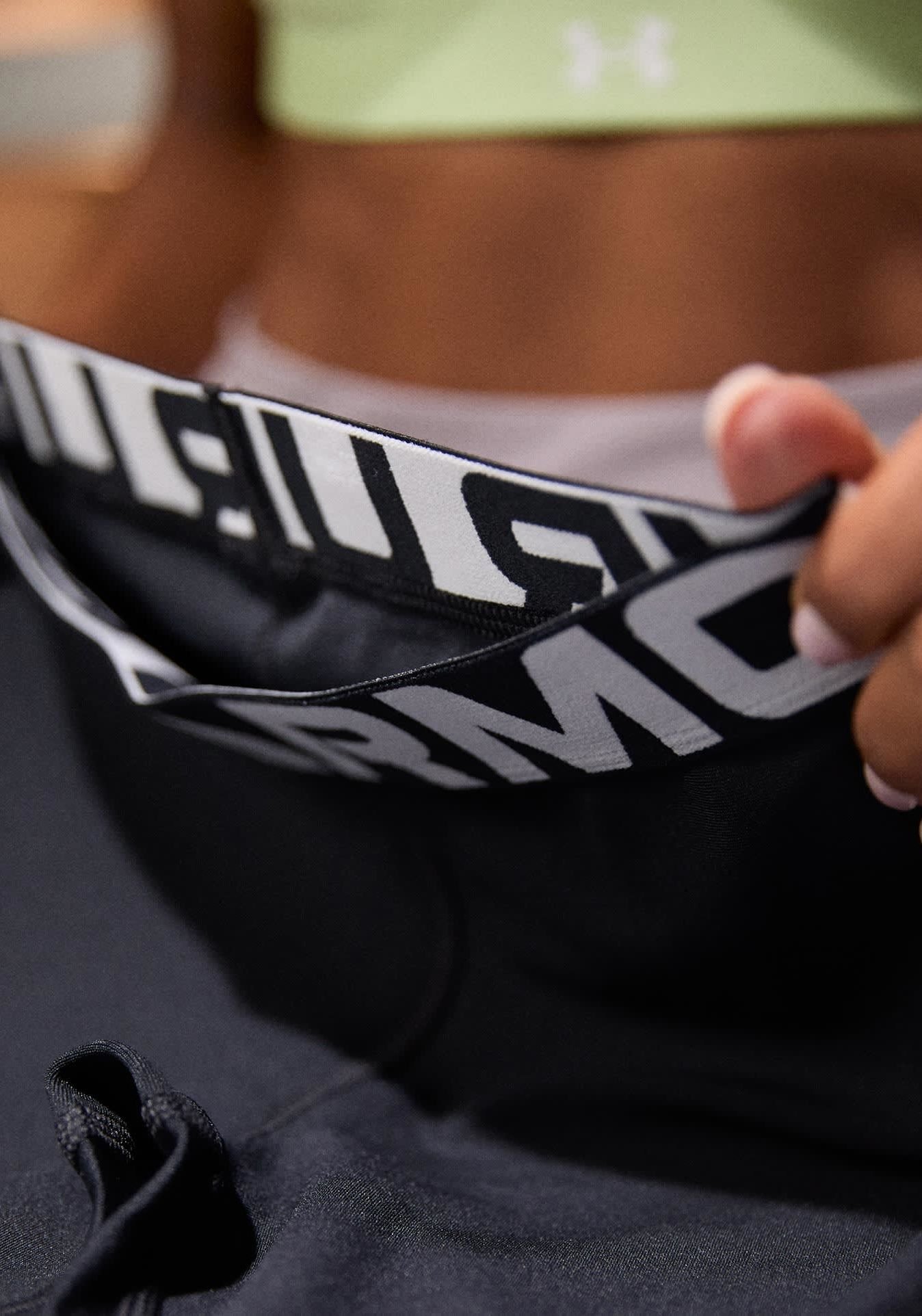 Schwarze Sportleggings mit einem weißen Markenbund. Der Bund trägt den Schriftzug "UNDER ARMOUR" in fetten Buchstaben. Texturiertes Material und Details mit Zugband sind sichtbar.