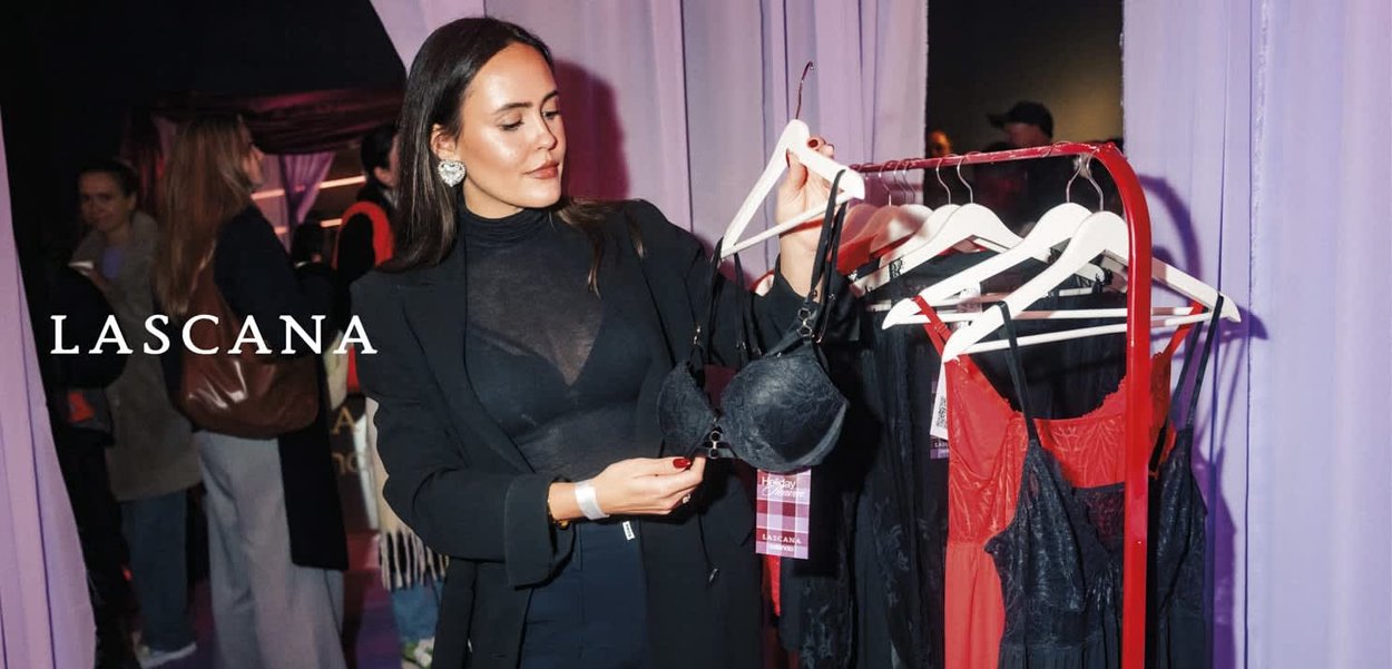 Kobieta w czarnej kurtce ogląda czarny biustonosz na czerwonym stojaku z różną bielizną w sklepie, w tle inni klienci.