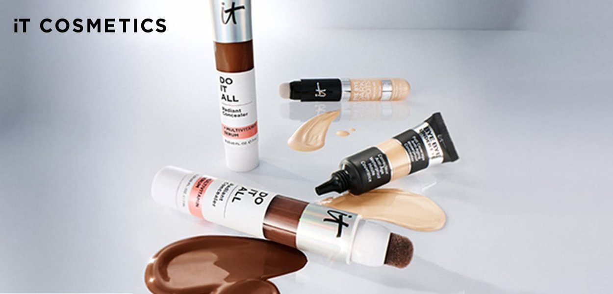 Verschiedene Farbtöne von IT Cosmetics Foundation- und Concealer-Tuben mit Produktmustern auf einer weißen Oberfläche.