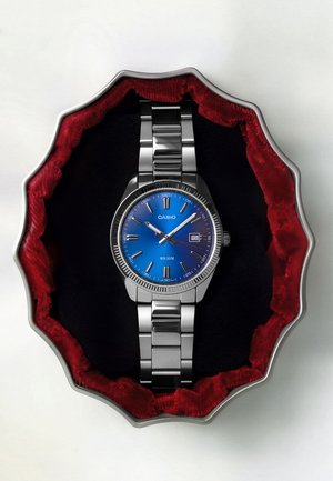 Zilverkleurig Casio polshorloge met blauwe wijzerplaat en datumaanduiding in een metalen kast met een rode fluwelen voering en kartelrand op een witte ondergrond.