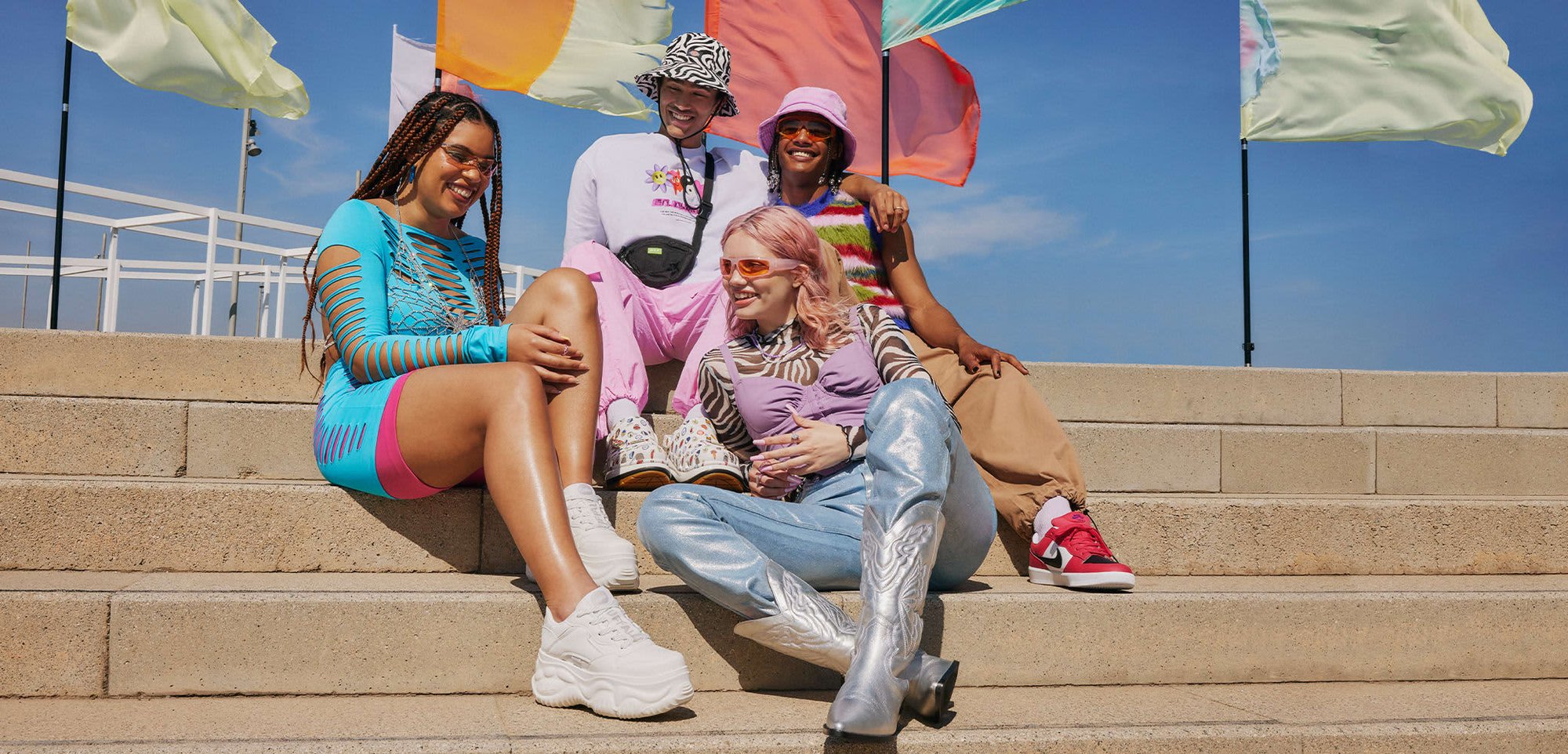 Groep van vier personen zittend op de trappen, gekleed in kleurrijke outfits: een blauwe jurk met uitsnijding, een gestreept shirt, metallic broek en gepatternde schoenen.