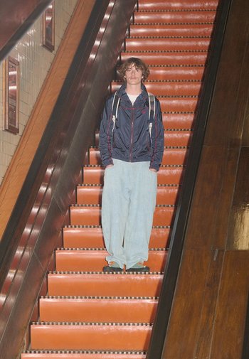 Een persoon draagt een marineblauwe jas, losse lichtblauwe jeans en zwarte schoenen terwijl hij op de oranje trap van een roltrap staat, met een houten leuning aan de zijkant.