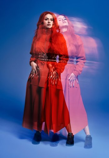 Rotes langärmliges Kleid mit hohem Kragen und geschichtetem Rock, tailliert mit einem silbernen Gürtel, getragen mit schwarzen Schuhen, vor einem blauen Hintergrund.